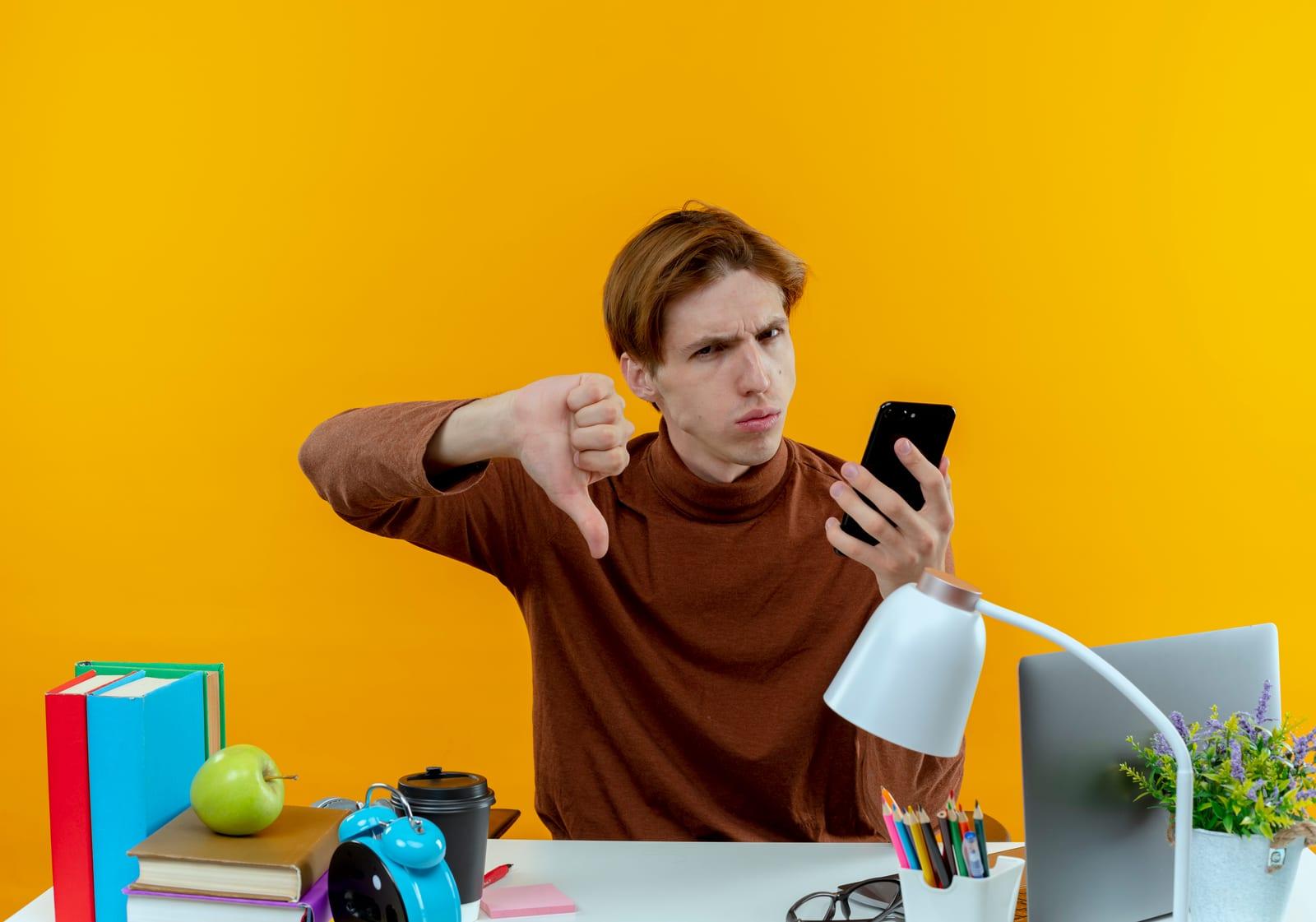 unpleased young student boy sitting at desk with school tools holding phone his thumb down isolated on yellow background